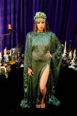 Woman in a green dress with a headpiece standing in front of a purple curtain with candles and decorative items.