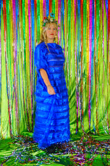 Model with blonde hair and a headdress wearing a RAZZLE DAZZLE tinsel tassel kaftan maxi dress with sheer mesh panels. The showstopping dress features shimmering tassels, a loose fit, and a slash neck. It can be worn free-flowing or cinched with hidden ribbons for a more fitted silhouette. Made from limited deadstock fabric, the semi-sheer dress is paired with just a bra and knickers in this photo, perfect for the festive season.