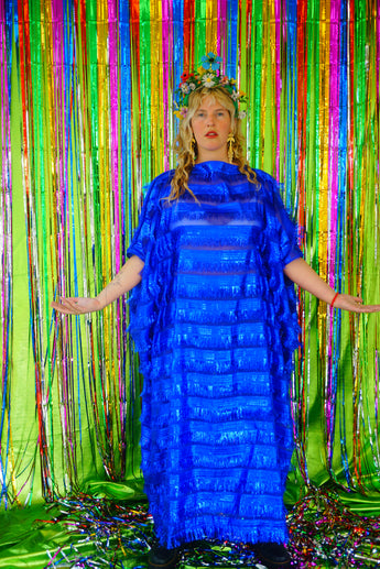 Model with blonde hair and a headdress wearing a RAZZLE DAZZLE tinsel tassel kaftan maxi dress with sheer mesh panels. The showstopping dress features shimmering tassels, a loose fit, and a slash neck. It can be worn free-flowing or cinched with hidden ribbons for a more fitted silhouette. Made from limited deadstock fabric, the semi-sheer dress is paired with just a bra and knickers in this photo, perfect for the festive season.