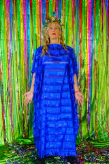 Front view of model with blonde hair and a headdress wearing a RAZZLE DAZZLE tinsel tassel kaftan maxi dress with sheer mesh panels. The showstopping dress features shimmering tassels, a loose fit, and a slash neck. It can be worn free-flowing or cinched with hidden ribbons for a more fitted silhouette. Made from limited deadstock fabric, the semi-sheer dress is paired with just a bra and knickers in this photo, perfect for the festive season.