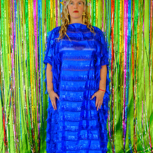 Model with blonde hair and a headdress wearing a RAZZLE DAZZLE tinsel tassel kaftan maxi dress with sheer mesh panels. The showstopping dress features shimmering tassels, a loose fit, and a slash neck. It can be worn free-flowing or cinched with hidden ribbons for a more fitted silhouette. Made from limited deadstock fabric, the semi-sheer dress is paired with just a bra and knickers in this photo, perfect for the festive season.