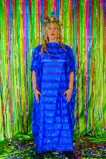 Model with blonde hair and a headdress wearing a RAZZLE DAZZLE tinsel tassel kaftan maxi dress with sheer mesh panels. The showstopping dress features shimmering tassels, a loose fit, and a slash neck. It can be worn free-flowing or cinched with hidden ribbons for a more fitted silhouette. Made from limited deadstock fabric, the semi-sheer dress is paired with just a bra and knickers in this photo, perfect for the festive season.