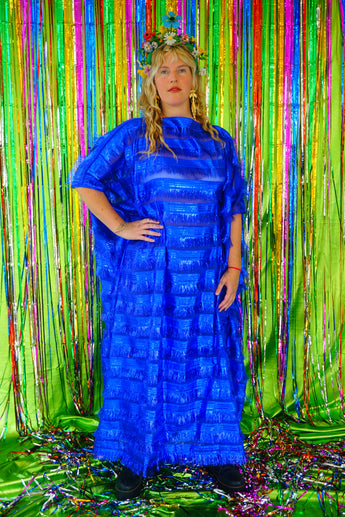 Model with blonde hair and a headdress wearing a RAZZLE DAZZLE tinsel tassel kaftan maxi dress with sheer mesh panels. The showstopping dress features shimmering tassels, a loose fit, and a slash neck. It can be worn free-flowing or cinched with hidden ribbons for a more fitted silhouette. Made from limited deadstock fabric, the semi-sheer dress is paired with just a bra and knickers in this photo, perfect for the festive season.