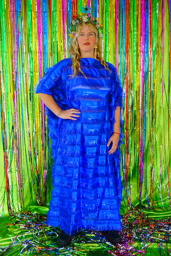 Model with blonde hair and a headdress wearing a RAZZLE DAZZLE tinsel tassel kaftan maxi dress with sheer mesh panels. The showstopping dress features shimmering tassels, a loose fit, and a slash neck. It can be worn free-flowing or cinched with hidden ribbons for a more fitted silhouette. Made from limited deadstock fabric, the semi-sheer dress is paired with just a bra and knickers in this photo, perfect for the festive season.