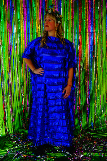 Model with blonde hair and a headdress wearing a RAZZLE DAZZLE tinsel tassel kaftan maxi dress with sheer mesh panels. The showstopping dress features shimmering tassels, a loose fit, and a slash neck. It can be worn free-flowing or cinched with hidden ribbons for a more fitted silhouette. Made from limited deadstock fabric, the semi-sheer dress is paired with just a bra and knickers in this photo, perfect for the festive season.