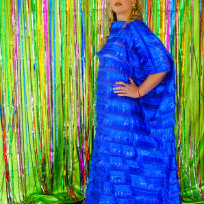 Side view of model with blonde hair and a headdress wearing a RAZZLE DAZZLE tinsel tassel kaftan maxi dress with sheer mesh panels. The showstopping dress features shimmering tassels, a loose fit, and a slash neck. It can be worn free-flowing or cinched with hidden ribbons for a more fitted silhouette. Made from limited deadstock fabric, the semi-sheer dress is paired with just a bra and knickers in this photo, perfect for the festive season.