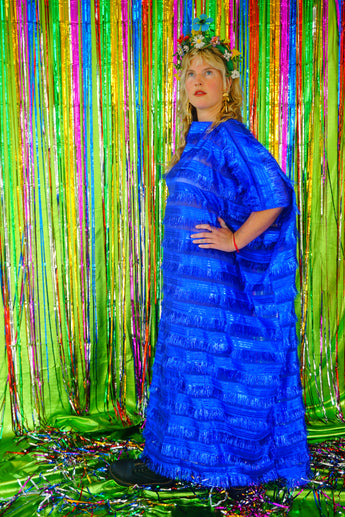 Side view of model with blonde hair and a headdress wearing a RAZZLE DAZZLE tinsel tassel kaftan maxi dress with sheer mesh panels. The showstopping dress features shimmering tassels, a loose fit, and a slash neck. It can be worn free-flowing or cinched with hidden ribbons for a more fitted silhouette. Made from limited deadstock fabric, the semi-sheer dress is paired with just a bra and knickers in this photo, perfect for the festive season.