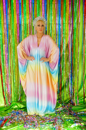 Model wearing a shimmering, liquid holographic maxi kaftan dress in pastel rainbow colors with a rianbow pompom headdress. 