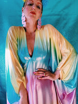 Close-up of model wearing a shimmering, liquid holographic maxi kaftan dress in pastel rainbow colors with a rainbow pompom headdress.