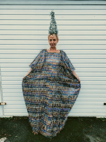 Front view of model wearing an iridescent maxi kaftan dress made from multi-colored shimmer tinsel in icy blues, whites, and bronzes, paired with a festive Christmas tree headdress. The dress features long, flowing tinsel strands that catch the light, creating a dazzling, holiday-inspired look. A special edition of the sellout tinsel maxi dress, perfect for the festive season.