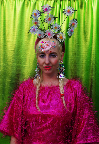 girl with blonde hair and plaits wears a pink shimmering tinsel mini dress with matching pink tights and heels against a metallic green backdrop