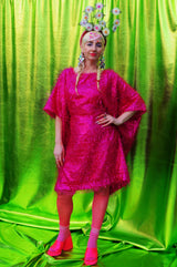 girl with blonde hair and plaits wears a pink shimmering tinsel mini dress with matching pink tights and heels against a metallic green backdrop