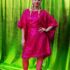 girl with blonde hair and plaits wears a pink shimmering tinsel mini dress with matching pink tights and heels against a metallic green backdrop