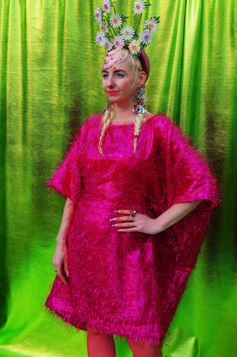 girl with blonde hair and plaits wears a pink shimmering tinsel mini dress with matching pink tights and heels against a metallic green backdrop