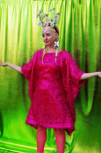 girl with blonde hair and plaits wears a pink shimmering tinsel mini dress with matching pink tights and heels against a metallic green backdrop