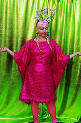 girl with blonde hair and plaits wears a pink shimmering tinsel mini dress with matching pink tights and heels against a metallic green backdrop