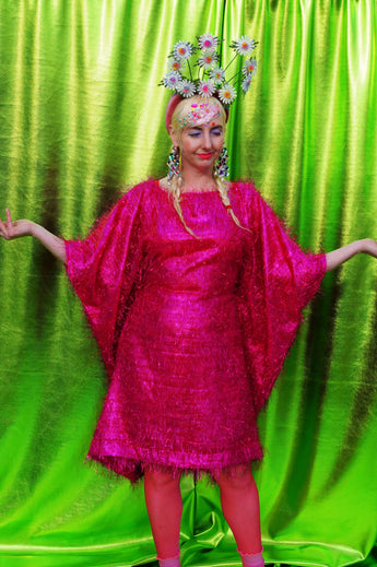 girl with blonde hair and plaits wears a pink shimmering tinsel mini dress with matching pink tights and heels against a metallic green backdrop