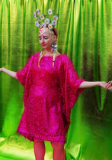 girl with blonde hair and plaits wears a pink shimmering tinsel mini dress with matching pink tights and heels against a metallic green backdrop