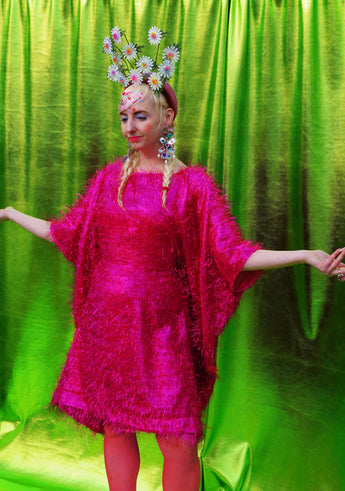 girl with blonde hair and plaits wears a pink shimmering tinsel mini dress with matching pink tights and heels against a metallic green backdrop