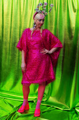 girl with blonde hair and plaits wears a pink shimmering tinsel mini dress with matching pink tights and heels against a metallic green backdrop