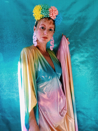 Close-up of model wearing a shimmering, liquid holographic maxi kaftan dress in pastel rainbow colors with a rainbow pompom headdress.