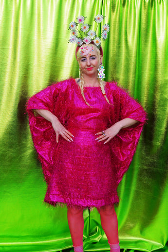 girl with blonde hair and plaits wears a pink shimmering tinsel mini dress with matching pink tights and heels against a metallic green backdrop