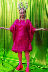 girl with blonde hair and plaits wears a pink shimmering tinsel mini dress with matching pink tights and heels against a metallic green backdrop