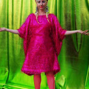 girl with blonde hair and plaits wears a pink shimmering tinsel mini dress with matching pink tights and heels against a metallic green backdrop