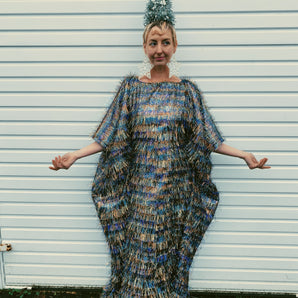Model wearing an iridescent maxi kaftan dress made from multi-colored shimmer tinsel in icy blues, whites, and bronzes, paired with a festive Christmas tree headdress. The dress features long, flowing tinsel strands that catch the light, creating a dazzling, holiday-inspired look. A special edition of the sellout tinsel maxi dress, perfect for the festive season.