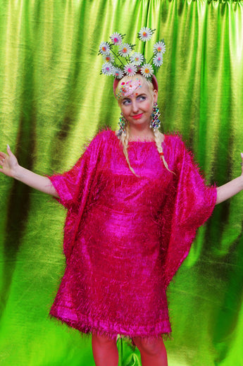 girl with blonde hair and plaits wears a pink shimmering tinsel mini dress with matching pink tights and heels against a metallic green backdrop