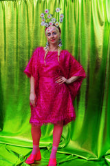 girl with blonde hair and plaits wears a pink shimmering tinsel mini dress with matching pink tights and heels against a metallic green backdrop