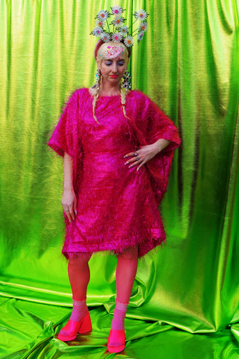 girl with blonde hair and plaits wears a pink shimmering tinsel mini dress with matching pink tights and heels against a metallic green backdrop