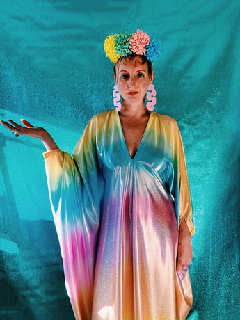 Close-up of model wearing a shimmering, liquid holographic maxi kaftan dress in pastel rainbow colors with a rainbow pompom headdress.