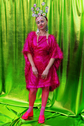 girl with blonde hair and plaits wears a pink shimmering tinsel mini dress with matching pink tights and heels against a metallic green backdrop