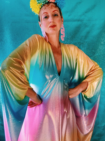 Close-up of model wearing a shimmering, liquid holographic maxi kaftan dress in pastel rainbow colors with a rianbow pompom headdress.