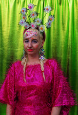 girl with blonde hair and plaits wears a pink shimmering tinsel mini dress with matching pink tights and heels against a metallic green backdrop