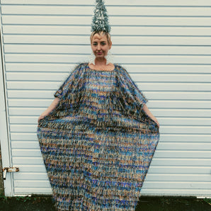 Model wearing an iridescent maxi kaftan dress made from multi-colored shimmer tinsel in icy blues, whites, and bronzes, paired with a festive Christmas tree headdress. The dress features long, flowing tinsel strands that catch the light, creating a dazzling, holiday-inspired look. A special edition of the sellout tinsel maxi dress, perfect for the festive season.