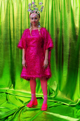 girl with blonde hair and plaits wears a pink shimmering tinsel mini dress with matching pink tights and heels against a metallic green backdrop