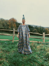 Model standing in a field wearing an iridescent maxi kaftan dress made from multi-colored shimmer tinsel in icy blues, whites, and bronzes, paired with a festive Christmas tree headdress. The dress features long, flowing tinsel strands that catch the light, creating a dazzling, holiday-inspired look. A special edition of the sellout tinsel maxi dress, perfect for the festive season.