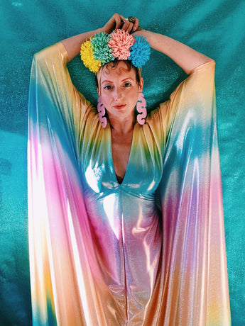 Close-up of model wearing a shimmering, liquid holographic maxi kaftan dress in pastel rainbow colors with a rianbow pompom headdress.