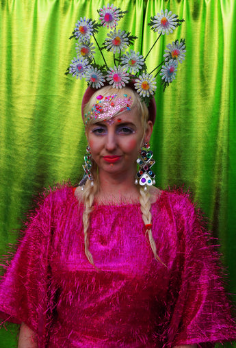 girl with blonde hair and plaits wears a pink shimmering tinsel mini dress with matching pink tights and heels against a metallic green backdrop