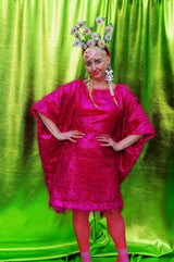 girl with blonde hair and plaits wears a pink shimmering tinsel mini dress with matching pink tights and heels against a metallic green backdrop