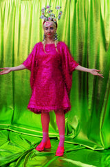 girl with blonde hair and plaits wears a pink shimmering tinsel mini dress with matching pink tights and heels against a metallic green backdrop